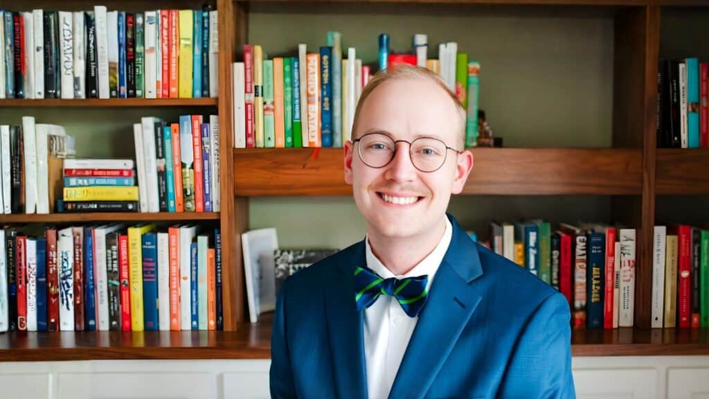 Young professional man in a blue suit and glasses smiling in front of a bookshelf.